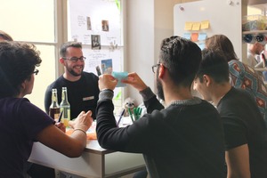 Fünf Menschen von hinten fotografiert. Sie stehen vor einem Tisch. Einer zeigt der Person, die hinter dem Tisch steht, einen Zettel.