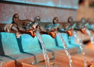 Reihe verzierter, patinierter Wasserhähne aus Metall, aus denen klares Wasser in Bögen fließt.