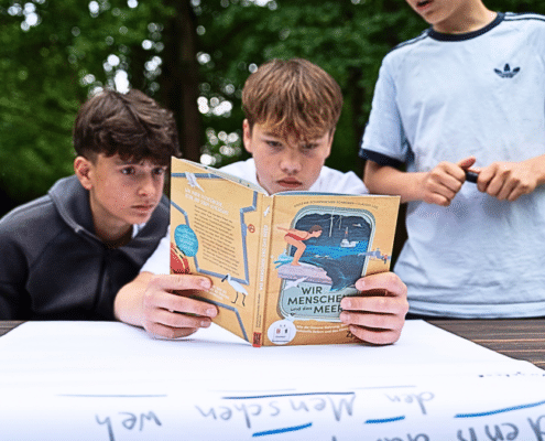 Das Editorial-Bild zeigt drei Schüler beim Testen eines Materials des Rucksack voll Meer. Hier begutachten sie das Buch Wir Menschen und das Meer.