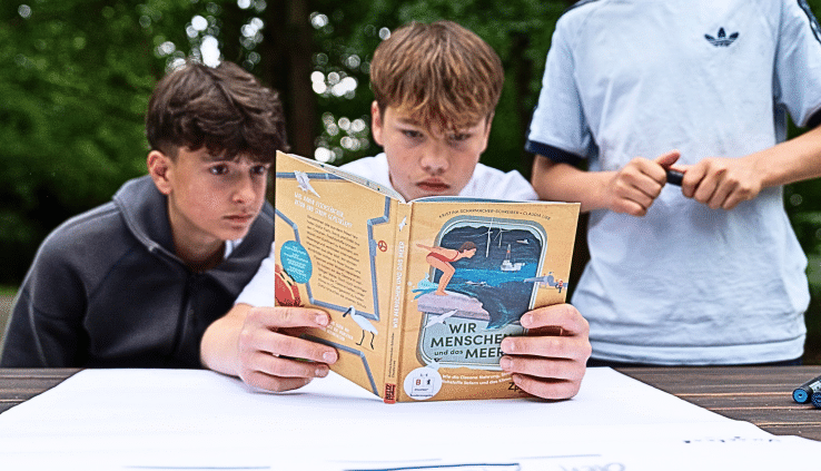 Das Editorial-Bild zeigt drei Schüler beim Testen eines Materials des Rucksack voll Meer. Hier begutachten sie das Buch Wir Menschen und das Meer.